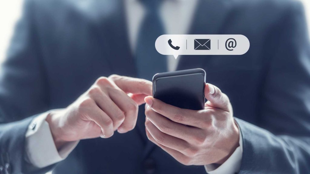 Frontalansicht eines Anzugträgers welcher ein Smartphone in den Händen haltet und Icons für tel, kontakt und @ erscheinen darüber